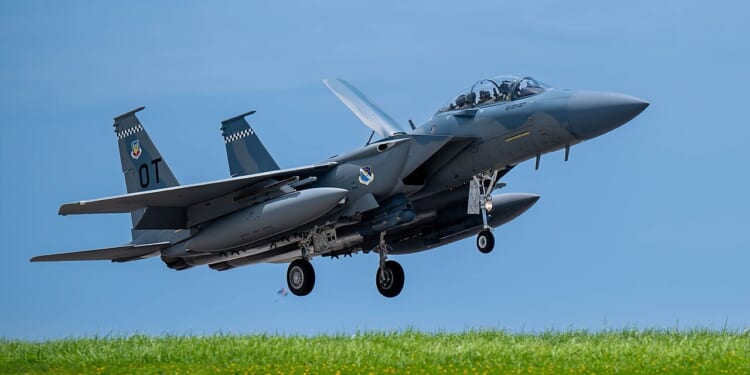 An F-15EX fighter jet taking off near a grassy hill.