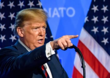 Donald Trump giving a speech at a NATO summit, in front of American flags.