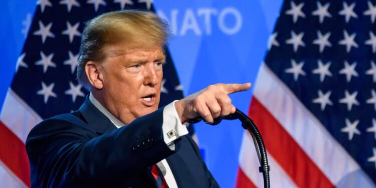 Donald Trump giving a speech at a NATO summit, in front of American flags.