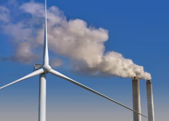 A wind turbine next to smokestacks from a fossil-fuel power plant.