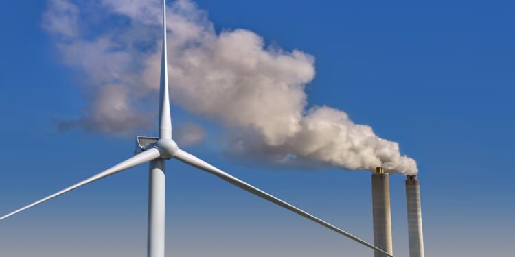 A wind turbine next to smokestacks from a fossil-fuel power plant.