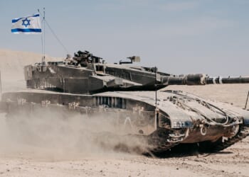 An tank in the desert flies an Israeli flag and kicks up dust.