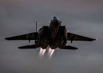 An F-15E Strike Eagle fighter jet in flight against a dark gray sky.